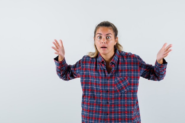 Foto gratuita giovane donna alzando le mani in camicia a quadri e guardando perplesso. vista frontale.