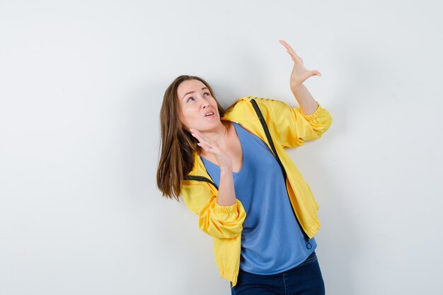 Young lady raising hands to defend herself in t-shirt, jacket and looking scared, front view.