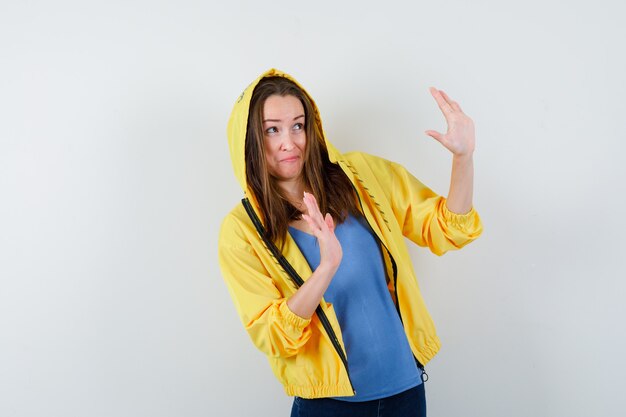 Young lady raising hands in protective manner in t-shirt, jacket and looking scared , front view.