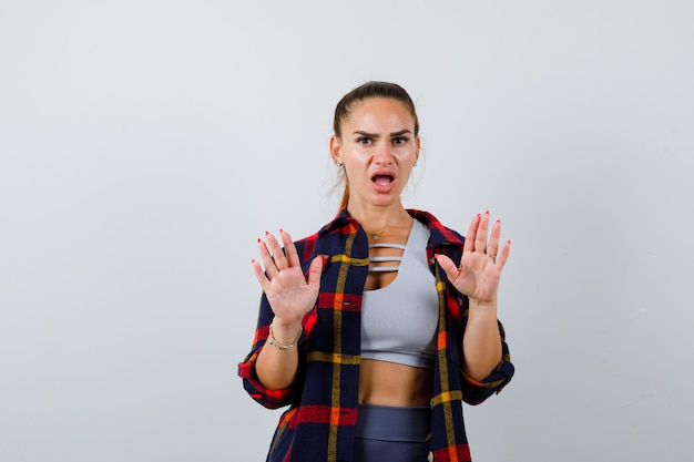 Free photo young lady raising hands in surrender gesture in top, plaid shirt and looking perplexed. front view.