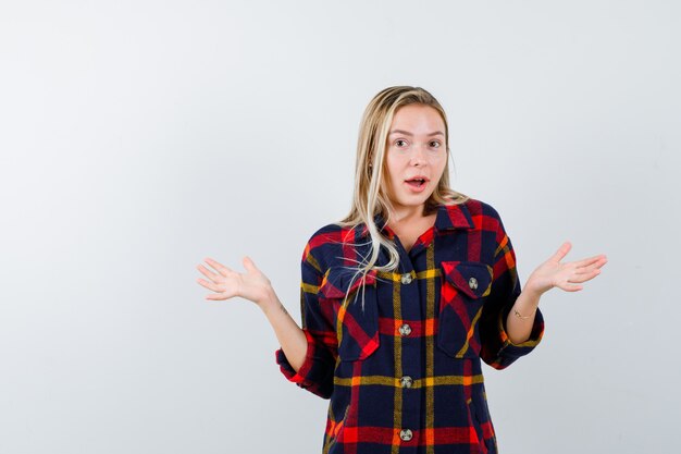 Young lady raising spread palms in checked shirt and looking puzzled , front view.