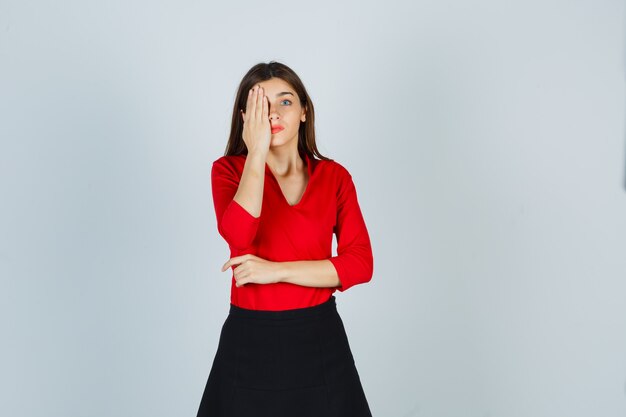 Young lady in red blouse, black skirt covering part of face with hand and looking pretty