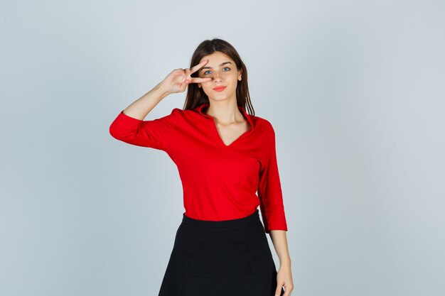 Young lady in red blouse, black skirt showing v sign on eye and looking confident