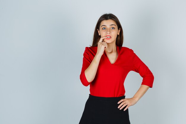 Young lady in red blouse, skirt biting finger while keeping hand on hip and looking scared