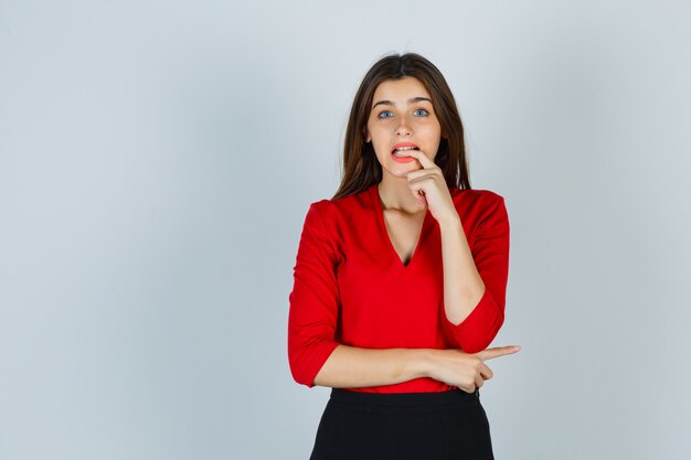 Young lady in red blouse, skirt biting finger while posing and looking forgetful