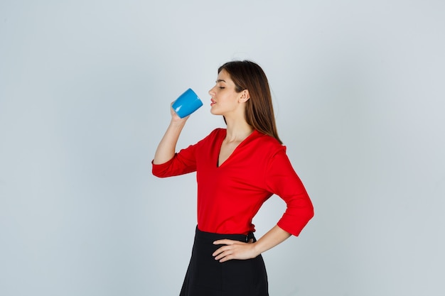 Young lady in red blouse, skirt drinking while keeping hand on hip and looking delighted