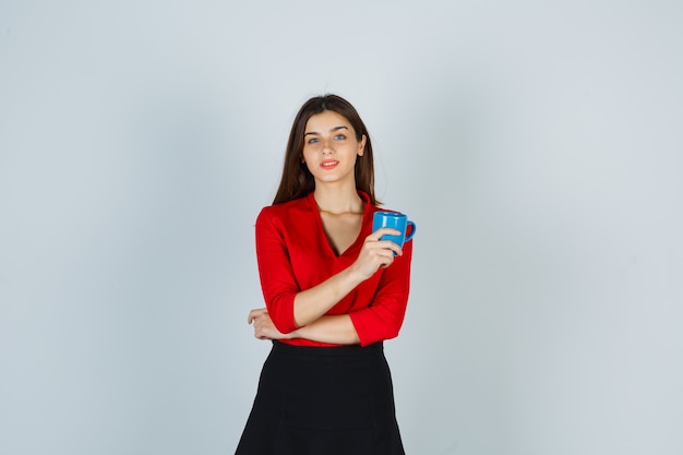 Young lady in red blouse, skirt holding cup while standing and looking charming