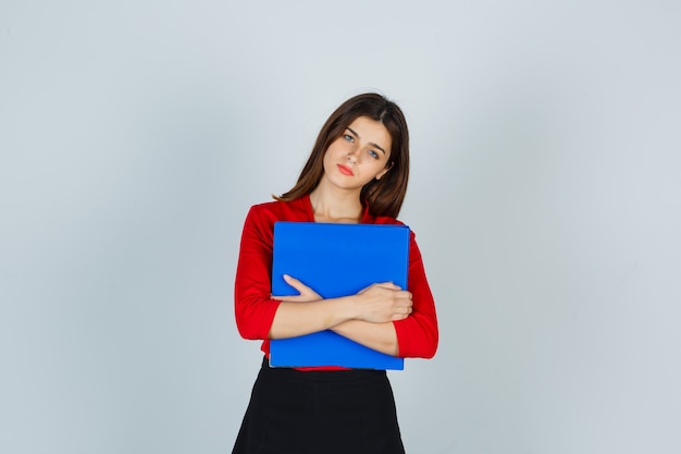 Young lady in red blouse, skirt holding folder on chest and looking pensive