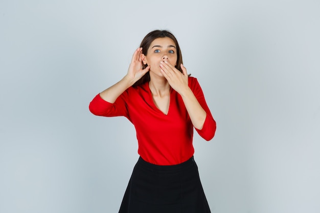 Young lady in red blouse, skirt holding hand behind ear while covering mouth