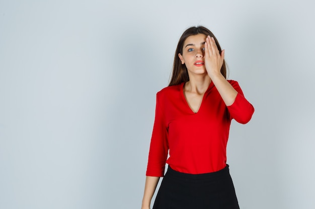 Young lady in red blouse, skirt holding hand on eye and looking serious