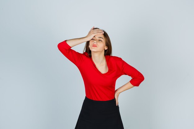 Young lady in red blouse, skirt holding hand on forehead while keeping hand