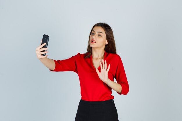 Young lady in red blouse, skirt making video call while waving hand