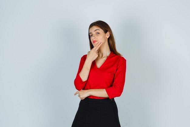 Young lady in red blouse, skirt posing while keeping fingers on chin and looking cute