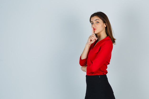 Young lady in red blouse, skirt showing silence gesture and looking serious
