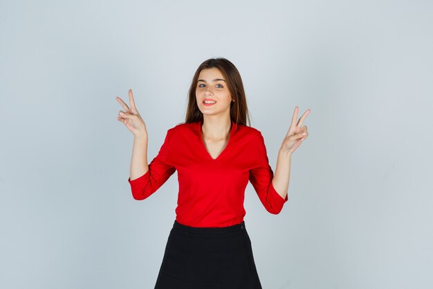 Молодая дама в красной блузке, юбке показывает знак победы и выглядит счастливой