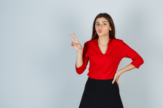 Young lady in red blouse, skirt showing victory sign while keeping hand on hip