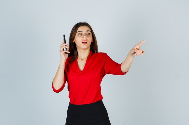 Young lady in red blouse, skirt talking on mobile phone while pointing aside