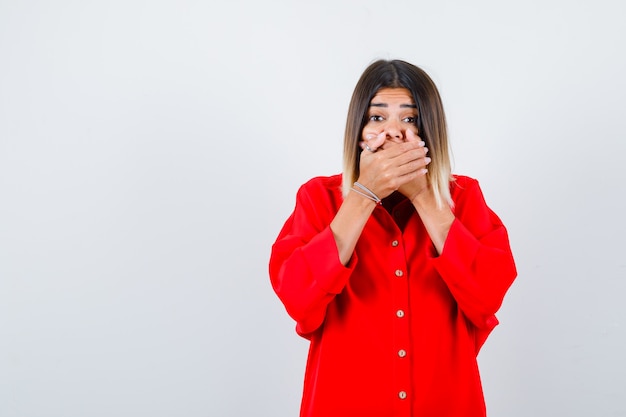 Young lady in red oversize shirt holding hands on mouth and looking puzzled , front view.