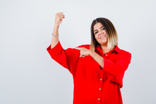 Young lady in red oversize shirt pointing at her muscles of arm and looking pleased , front view.