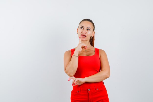 Young lady in red singlet, red trousers holding finger on chin and looking perplexed , front view.