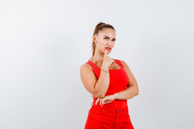 Young lady in red singlet, red trousers holding finger on mouth and looking pensive , front view.