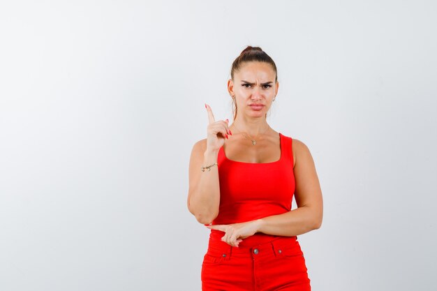 Young lady in red singlet, red trousers pointing up and looking serious , front view.