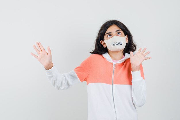 Young lady rejecting something with her open palms in hoodie, face mask and looking anxious.