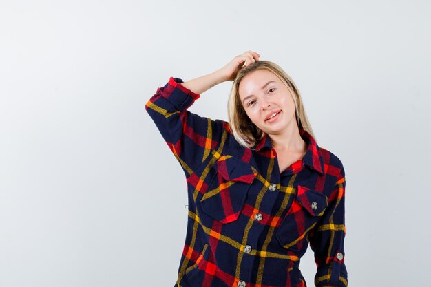 Giovane donna che graffia la testa in camicia a quadri e sembra affascinante, vista frontale.