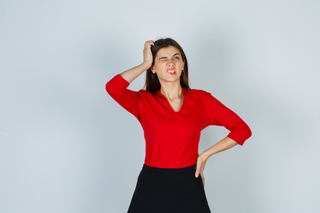 Young lady scratching head while keeping hand on hip in red blouse