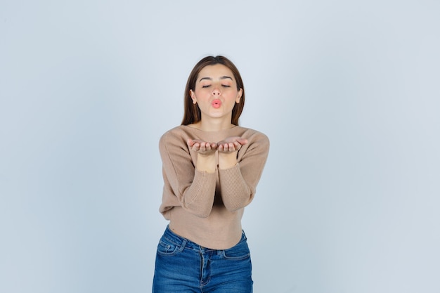 Young lady sending air kiss in beige sweater, jeans and looking pretty. front view.