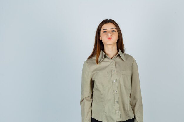 Young lady in shirt blowing cheeks, pouting lips and looking puzzled , front view.