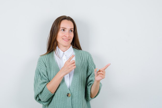 Young lady in shirt, cardigan pointing away and looking pleased , front view.