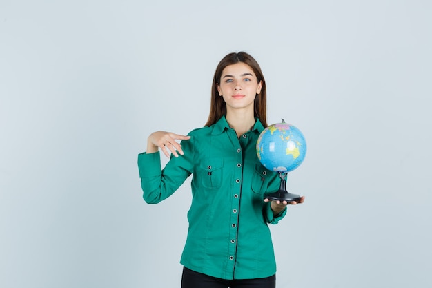 Free photo young lady in shirt holding earth globe, pointing aside and looking pleased , front view.