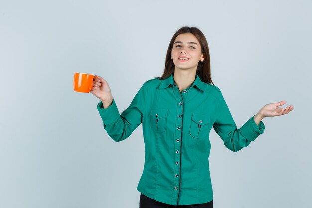 Молодая дама в рубашке держит оранжевую чашку чая, разводит ладонь в сторону и выглядит весело, вид спереди.