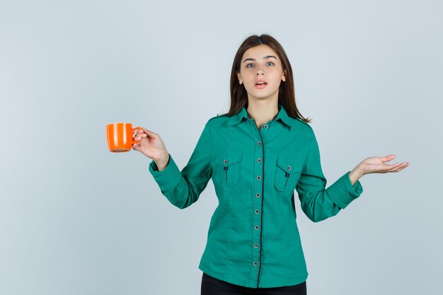 Young lady in shirt holding orange cup of tea, spreading palm aside and looking focused , front view.