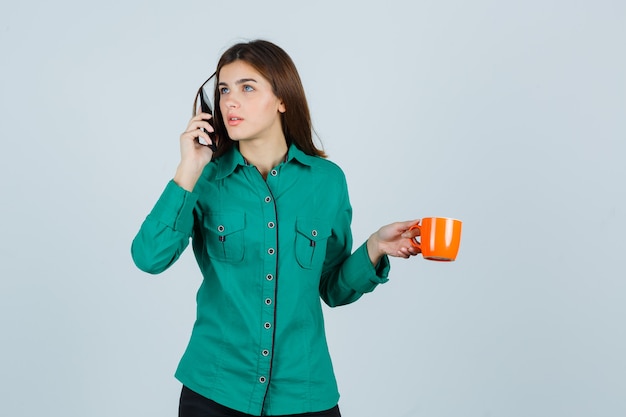 Young lady in shirt holding orange cup of tea, talking on the mobile phone and looking careful , front view.