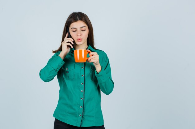 Young lady in shirt holding orange cup of tea, talking on the mobile phone and looking confident , front view.