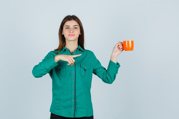 右側を指して、自信を持って、正面図を見て、オレンジ色のお茶を持っているシャツを着た若い女性。