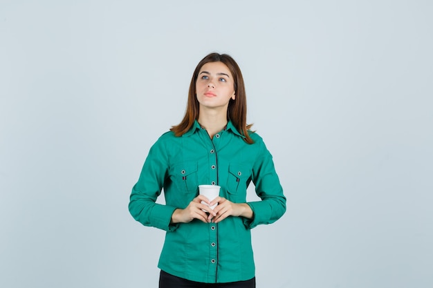 Молодая дама в рубашке держит пластиковую чашку кофе и смотрит задумчиво, вид спереди.