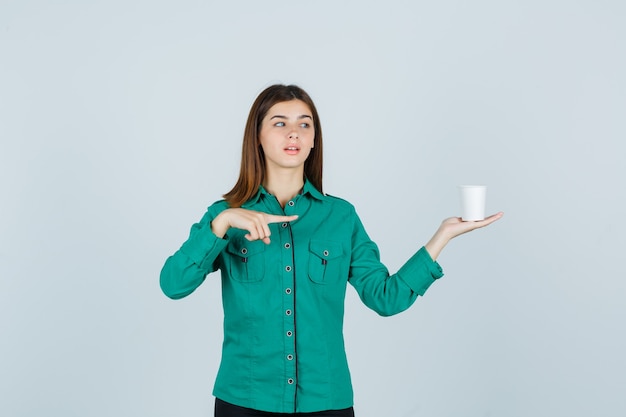 Young lady in shirt holding plastic cup of coffee while pointing to the right side and looking focused , front view.