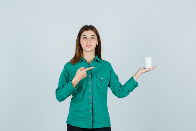 Young lady in shirt holding plastic cup of coffee while pointing to the right side and looking focused , front view.