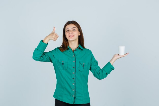 Young lady in shirt holding plastic cup of coffee while showing thumb up and looking cheery , front view.