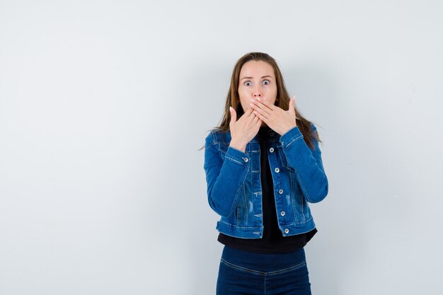 Young lady in shirt, jacket holding hands on mouth and looking shocked , front view.