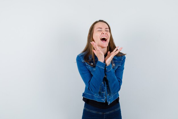 Young lady in shirt, jacket raising hands while screaming and looking happy , front view.