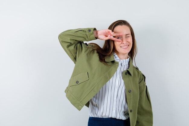 Young lady in shirt, jacket showing V-sign on eye and looking confident , front view.