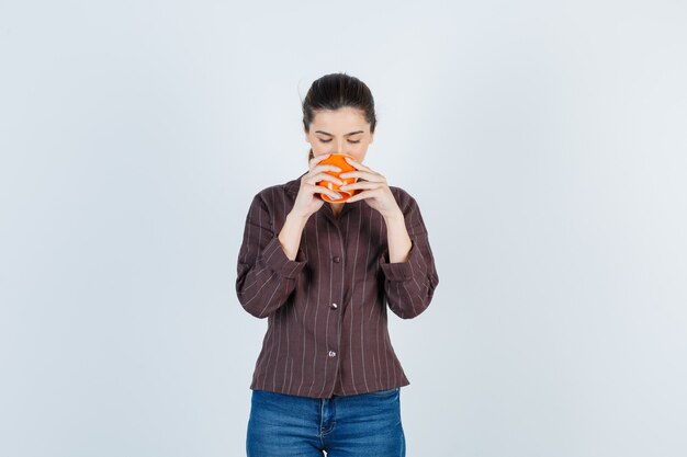 Young lady in shirt, jeans drinking from cup and looking pleased , front view.