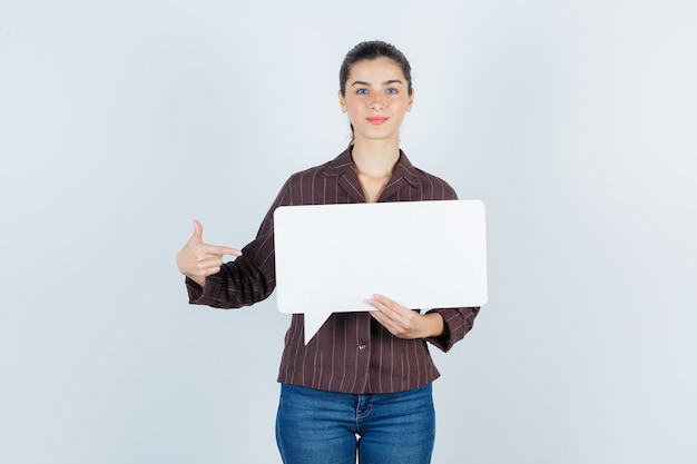 Young lady in shirt, jeans pointing to the side, keeping paper poster and looking satisfied , front view.