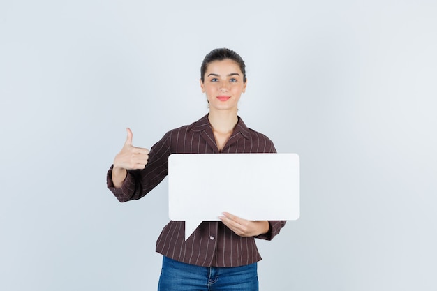 Young lady in shirt, jeans showing thumb up, keeping paper poster and looking pleased , front view.