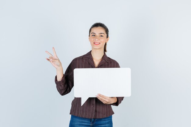Young lady in shirt, jeans showing V-sign, keeping paper poster and looking happy , front view.