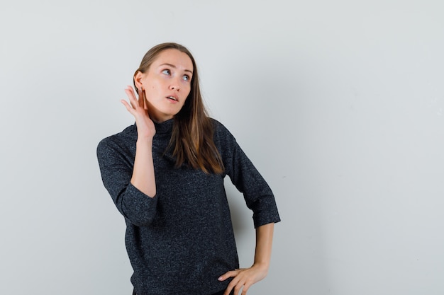 Young lady in shirt keeping hand behind ear and looking curious